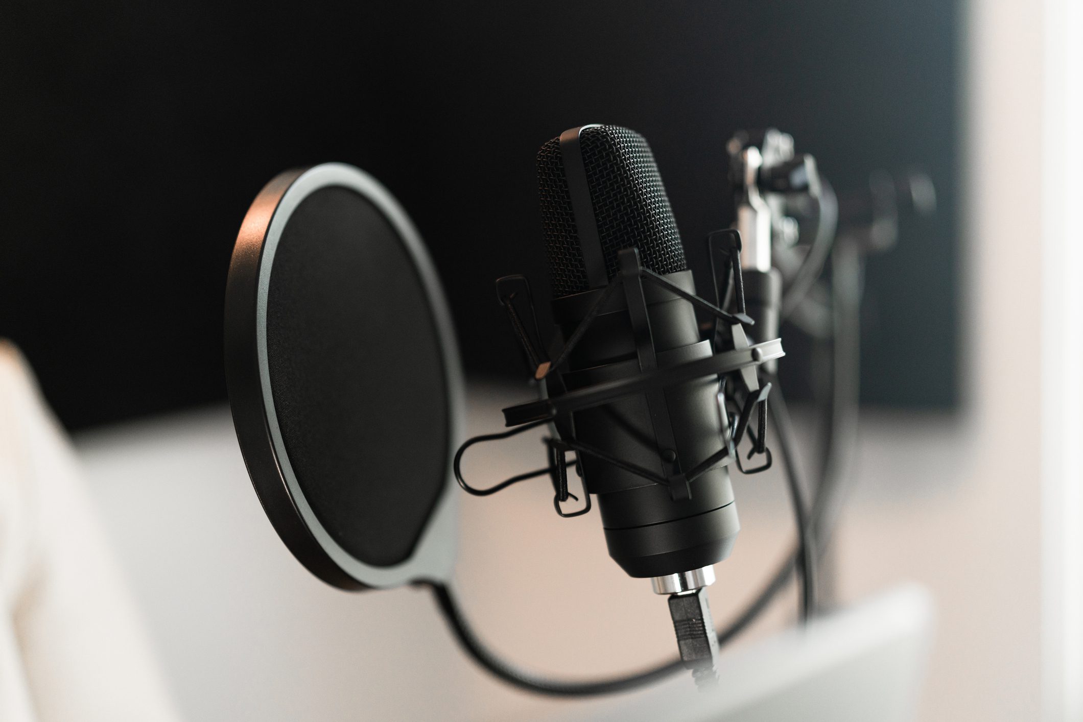 Close-up of a black condenser microphone with a pop filter, positioned on a desk stand in a recording studio setting.