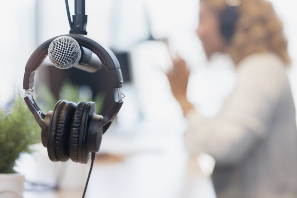 Close-up of a microphone and headphones hanging in a recording studio, with a person wearing headphones speaking in the blurred background.