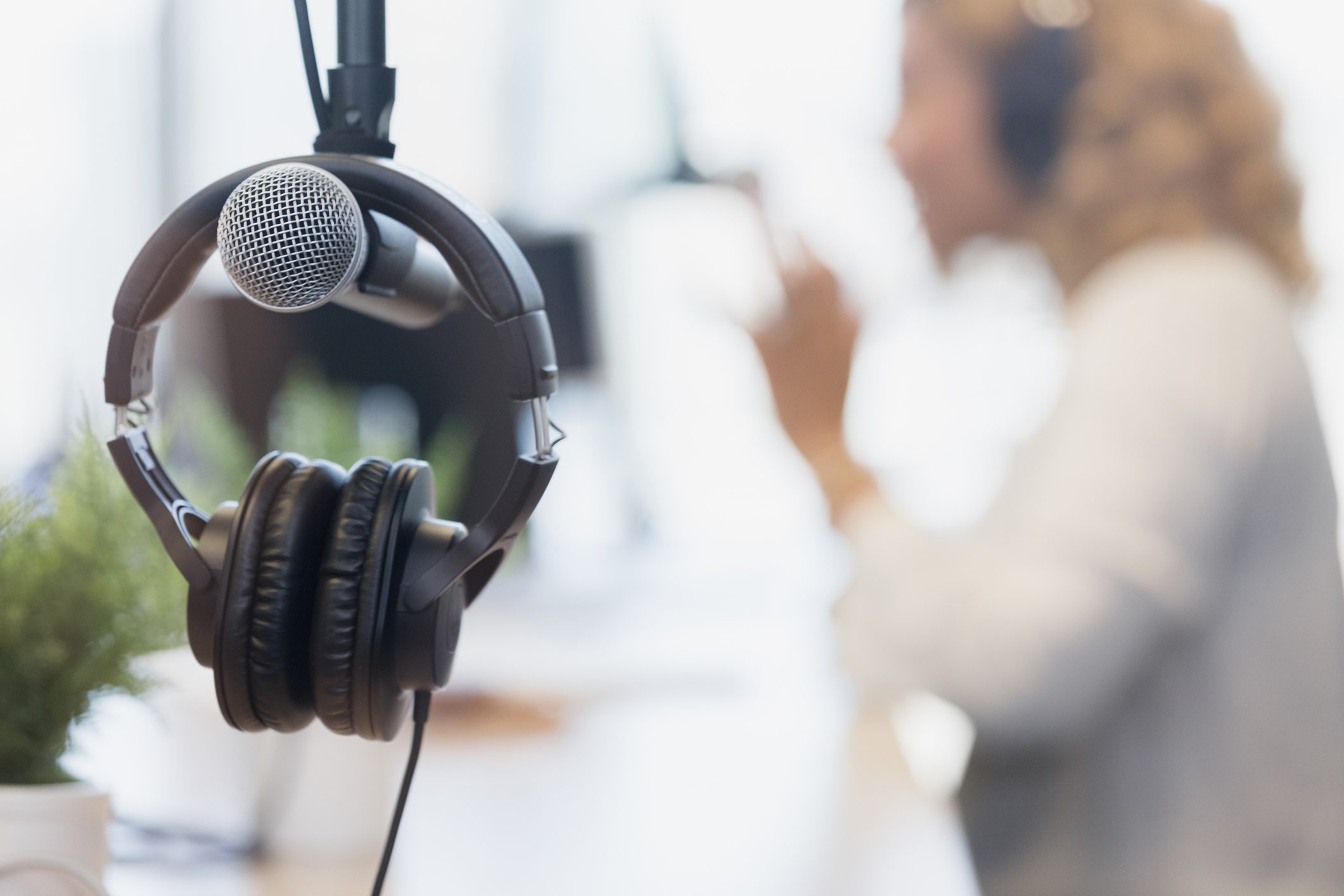 Close-up of a microphone and headphones hanging in a recording studio, with a person wearing headphones speaking in the blurred background.
