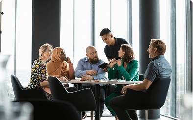 Six people sit and stand around a table in a modern office, engaged in discussion with documents and a tablet in hand. Large windows provide natural light.