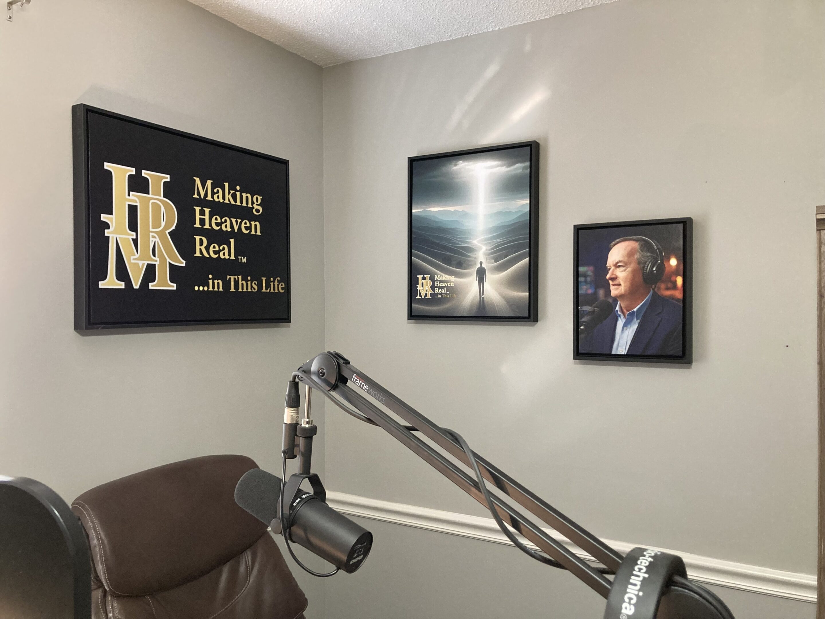A podcast recording setup with a microphone arm and three framed wall art pieces, including text and portraits, displayed on a light gray wall.