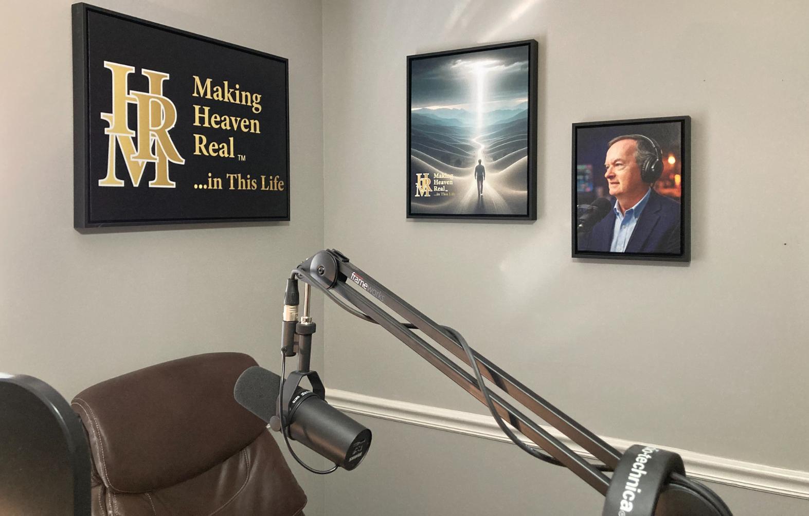 A podcast recording setup with a microphone arm, chair, and three framed wall art pieces, including a portrait and motivational text, in a neutral-colored room.