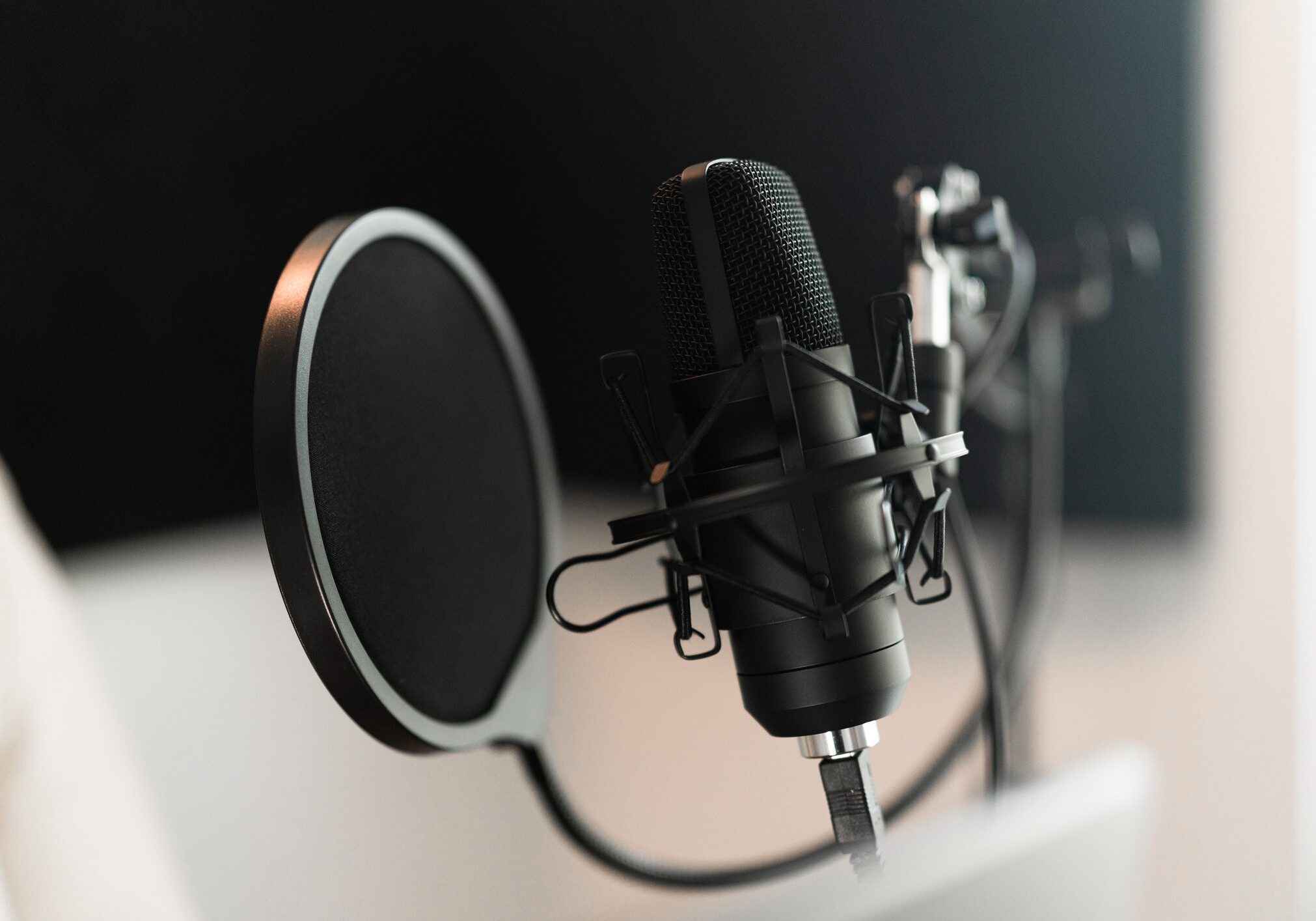 Close-up of a black condenser microphone with a pop filter, positioned on a desk stand in a recording studio setting.