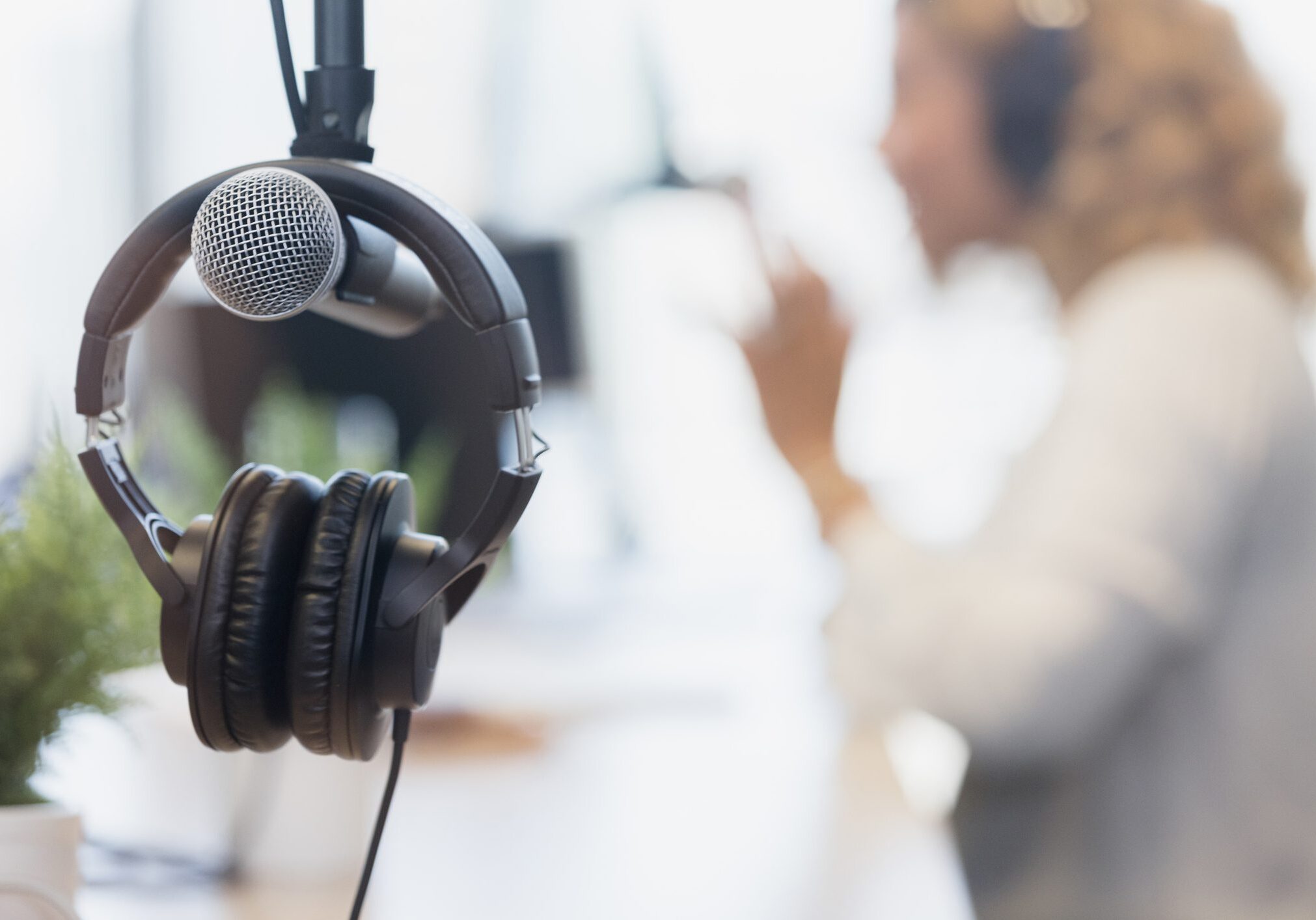 Close-up of a microphone and headphones hanging in a recording studio, with a person wearing headphones speaking in the blurred background.